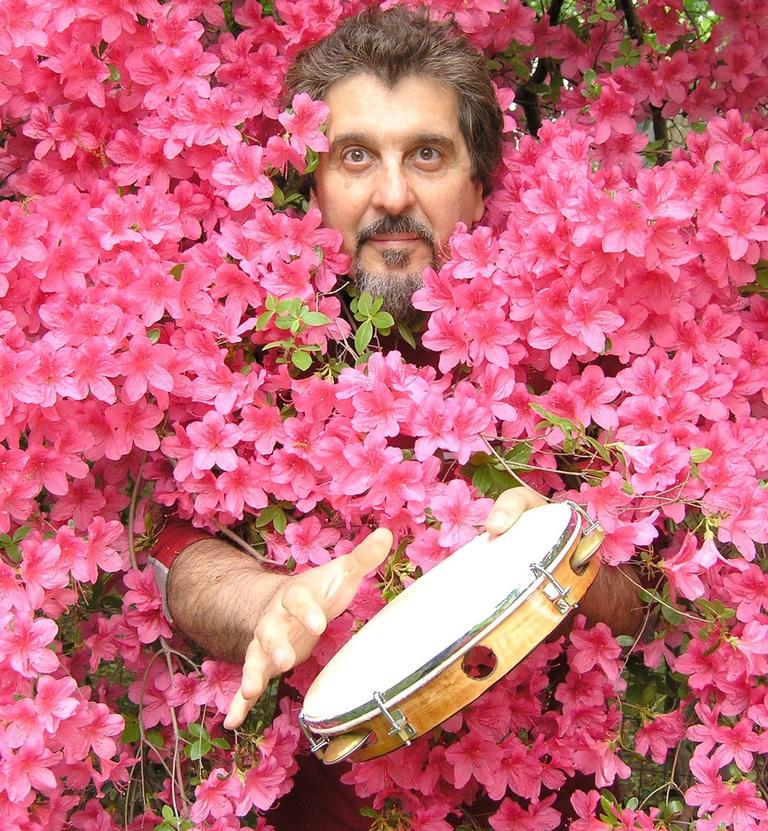  A man’s face and outstretched hands emerge from a wall of pink flowers. He is playing a tambourine. 