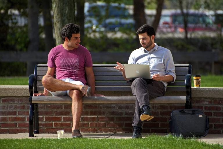 Two men sitting on a park bench. One is talking to the other with a laptop on his lap. It's a sunny warm day.
