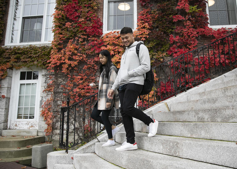 Two students walking down the steps outside of a campus building. It is fall and the ivy on the side of the building is changing color.