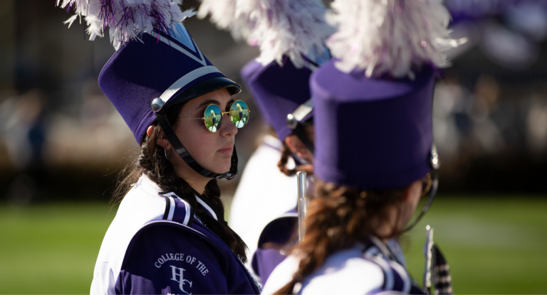 Band students dressed in band garb. They are wearing purple hats with a feather on top and seem to be waiting for their que to play.