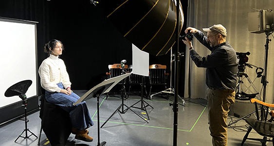 young womans sitting in lamplight, male photographer at right pointing camera in studio setting