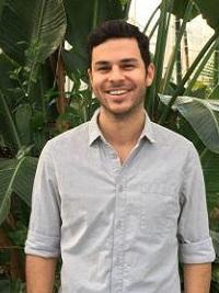 faculty member with dark hair and dark facial hair smiling at the camera with large plants behind him