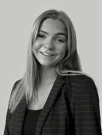 In this black and white headshot, Sasha, who has blond hair and is wearing a blazer, looks at the camera and smiles.