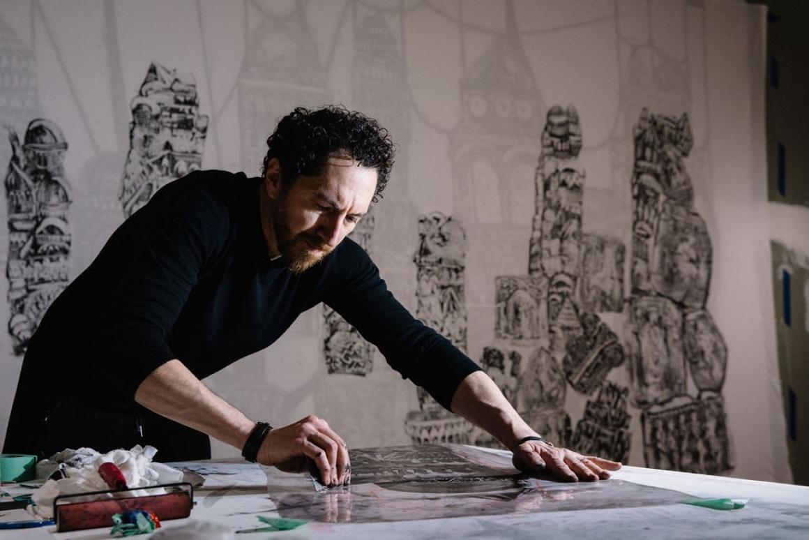 Kevork Mourad bends over a table, drawing. Images of his work in black and white are visible in the background.