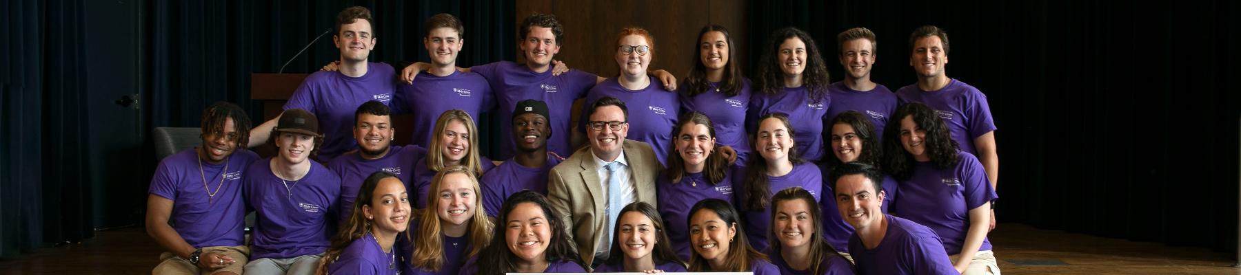 Group of 26 smiling students with purple Holy Cross shirts and khaki shorts/pants on posing for a picture