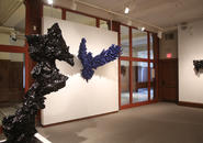 Large black abstract sculpture of a horse in foreground, large blue winged sculpture on wall in background