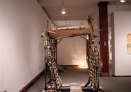 Gate constructed of hockey sticks, branches, and woven materials in foreground, swirling light sculpture in background, 