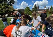 A candid shot of about 11 students gathered around a table outside. They are doing a craft: a foil pan with a white substance and they are adding red and blue food coloring. It is a sunny day with big clouds in the sky.