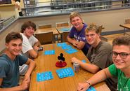 Five students gathered around a table, smiling and looking at the camera. They are playing bingo in a hall near a dining area.