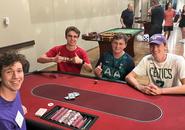 Four students at a gambling table. One student is the dealer with lots of chips in front of him. Two students have a smaller amount of chips and one student appears to have no ships. All students are smiling and one student is giving two thumbs up.