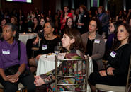 A large group of women sitting at tables and standing in the back of the room listening to a speaker at an event.
