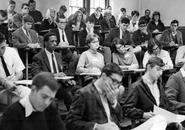 A black and white photo from the 60's of students in a lecture hall. While predominantly male, there are about 8 women out of a total of about 31 students