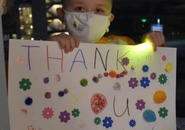 A young bald girl named Lily holds a sign that reads "thank you - love, Lily" that is covered in stickers.
