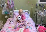 A young bald girl is pictured smiling in the hospital bed and room decorated with pink and unicorns.