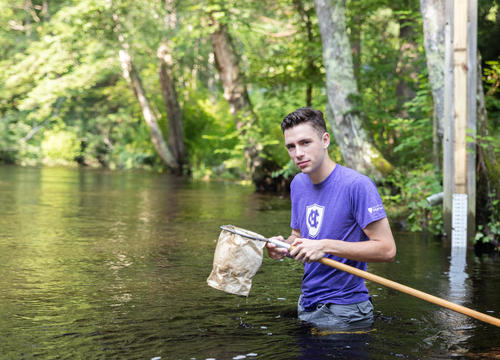 A student in a Holy Cross shirt stands in the Blackstone River with a bug catching net