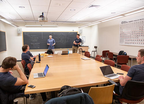 Students sit around a large table while one stands at a blackboard at the front of the room