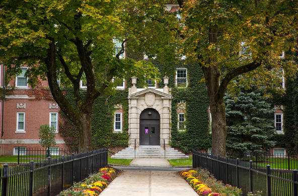 Entrance to a residence hall as framed by trees and rows of chrysanthemums. 