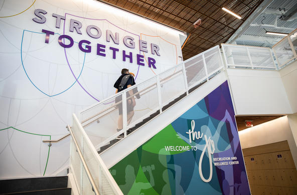 A student walking up the stairs inside at the Jo Recreation and Wellness Center. The wall adjacent to the stairs is white with an outline of the Holy Cross shield stenciled in various colors and the word Stronger Together span a decent surface on the wall.