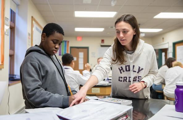 A Holy Cross student helping a middle school student with a school assignment. The student is pointing to something on a sheet of paper and the middle schooler, is listening to her and looking at what she is pointing at while holding a pencil.
