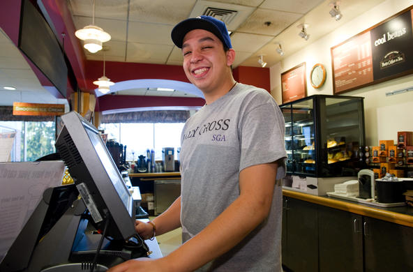 Student working in a cafeteria. He is wearing a hat and a gray Holy Cross tshirt. He is smiling and looking down at the camera from the side and is standing in front of a cash register.