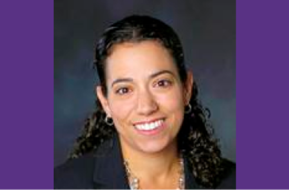 A picture of Professor Lauren Capotsoto. She has long, dark curly hair pulled half up. She is wearing a dark blazer and a light colored button down shirt. She is smiling.