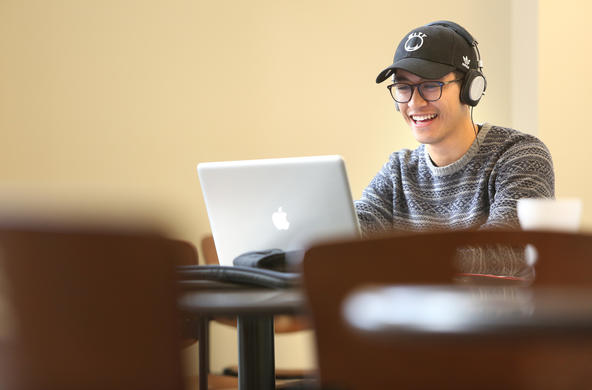 A male students with headphones on working at his mac laptop. He is wearing a black baseball cap, black rimmed glasses, and a gray and black striped long sleeve shirt.
