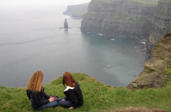 two students sitting on the grass on a cliff overlooking water