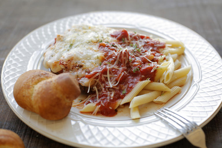 A plate of chicken parm, ziti and a garlic knot with a fork resting on the side of the plate. 