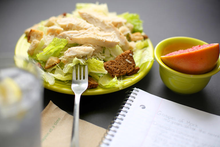Plate of chicken caesar salad, small bowl with half a grapefruit, ice water with wedge of lemon, napkin and notebook. 