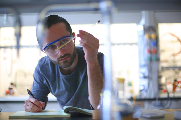 student in a lab wearing goggles and looking at a long tube and taking notes in a notebook