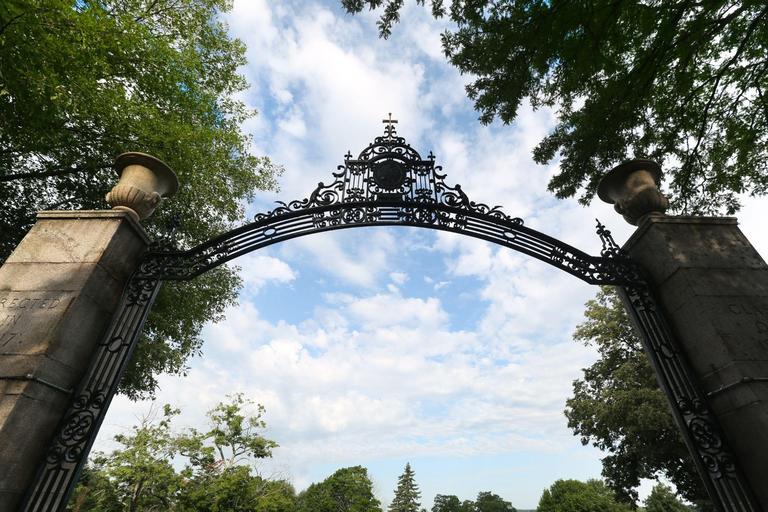 picture of the front gate looking up from underneath, blue sky with some clouds, green trees on each site with two stone columns and black iron archway