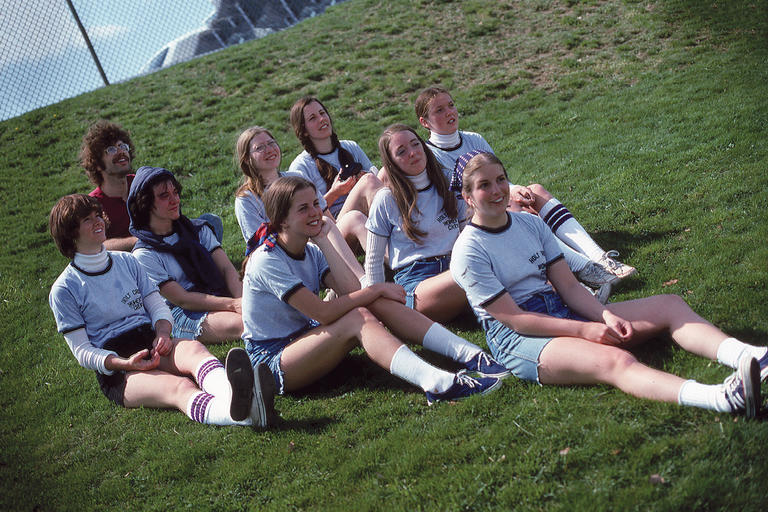 A color photo of 8 women and one man dressed in Holy Cross crew shirts. They are women are sitting on the grass looking presumably at another camera taking a similar photo.