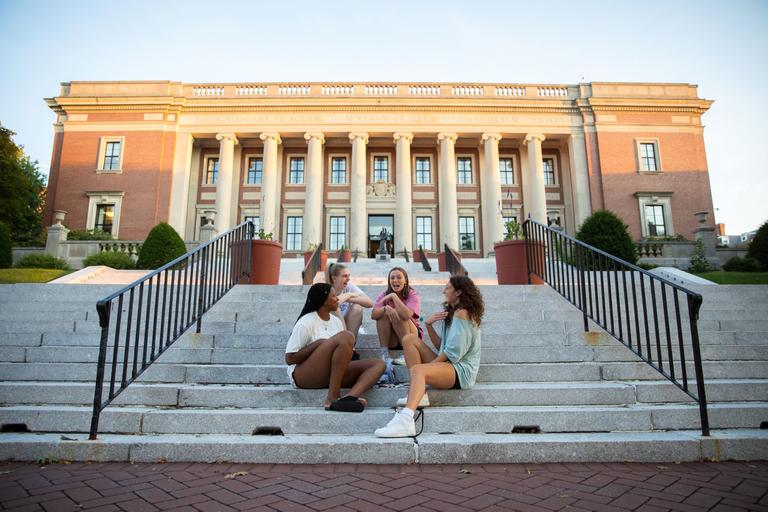 A group of several students sitting on the steps leading up to a campus on building. They are all engaged in a conversation.