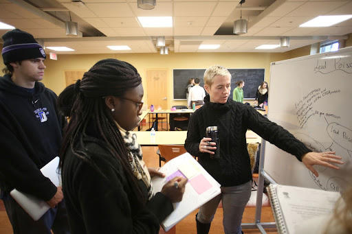 An instructor working with two students at a whiteboard. The students are holding a clipboard and one student is taking notes while the instructor, who is holding a cup of coffee/tea/water is pointing at the whiteboard.