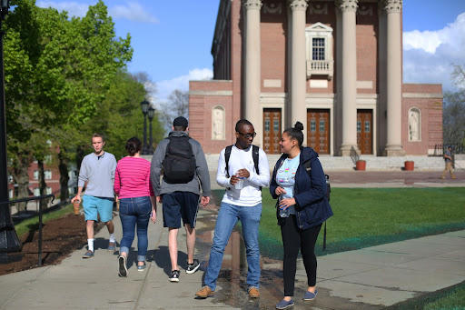 Students outside on campus on a sunny day. Three students are walking away from the camera and two students are walking toward the camera talking to each other.