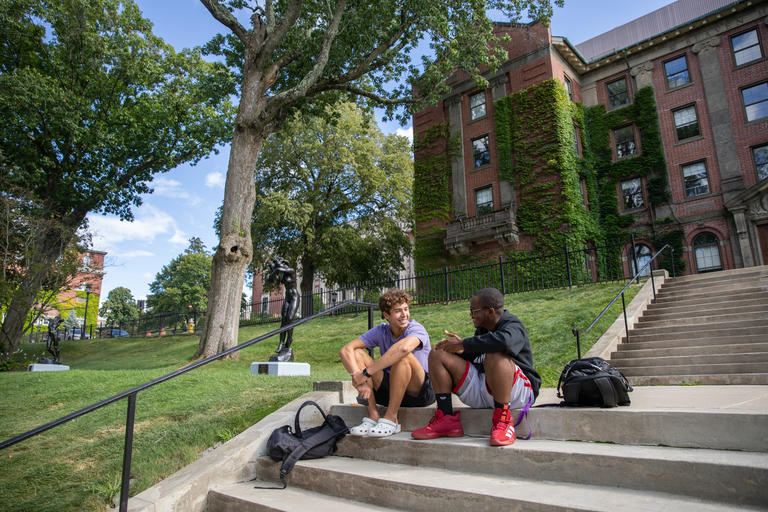 Two students sitting on stairs leading up to a campus building talking. The weather is warm and sunny and the young men are smiling.