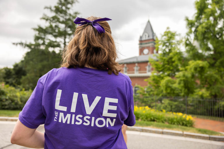 Student posing with her back to the camera. She is wearing a Holy Cross purple tshirt with the words Live the Mission. Her hair is half up, half down and she wears a purple ribbon.