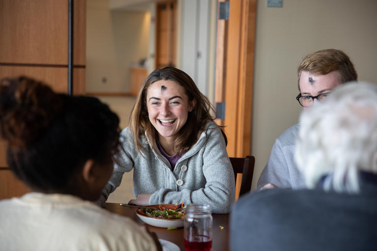 Photo of a student smiling with an ash Wednesday smudge. She is at a retreat and talking to another student across the table from her while they enjoy a meal.