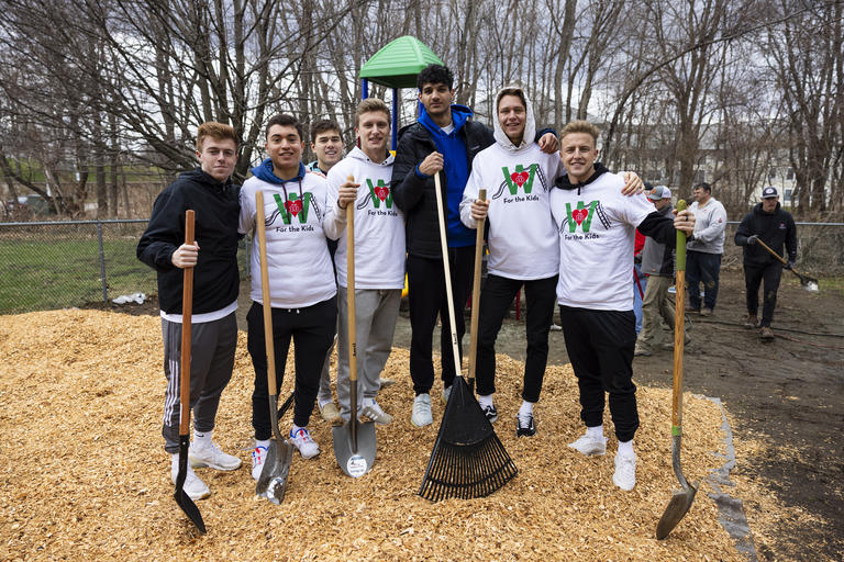 A group of 7 students standing on a playground. Some have shovels in their hands and one has a rake. They are laying down fresh mulch at a community event.