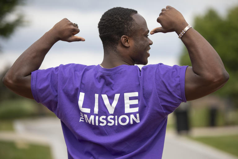 A man with his back to the camera wearing a Holy Cross purple shirt that says "Live the Mission". Josh is pointing to the back of his shirt with both thumbs.
