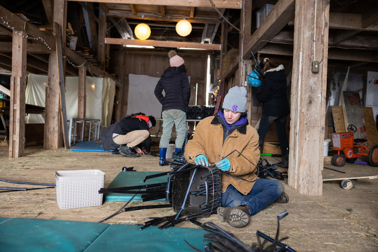 Three students in a barn at Sun Moon Farm. One student is sitting on the floor wrapping a bundle of plastic ties and the other two are in the background with their backs to the camera. 