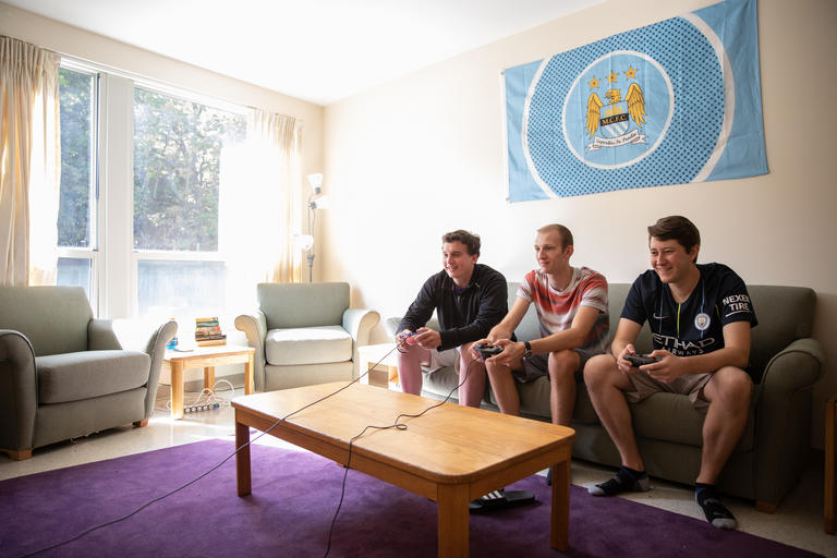 3 students in a residential hall, playing video games and smiling. The sun is shining through the window and the students are having a good time.