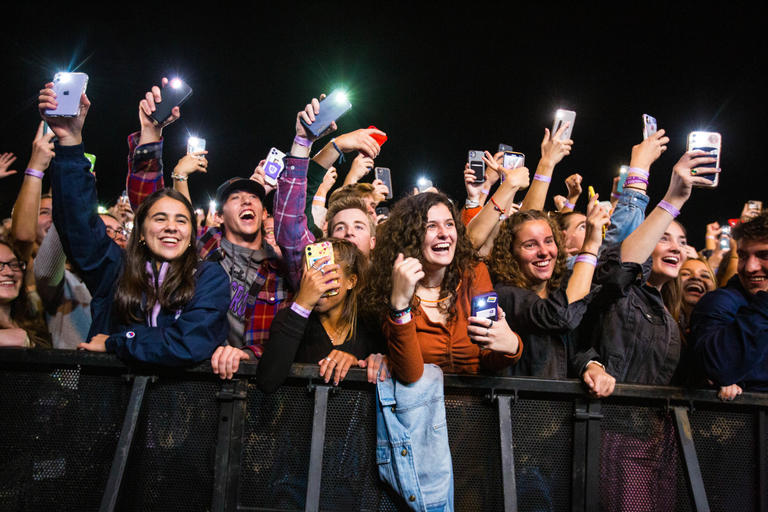 A group of students at a concert. It is nighttime and all the students are smiling and standing. Most are holding up their mobile phones and have the flashlight function on.