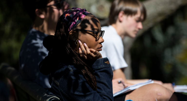 Students sitting outside partially in the shade with notebooks on their laps and pencils in their hands listening to someone who is not pictured.