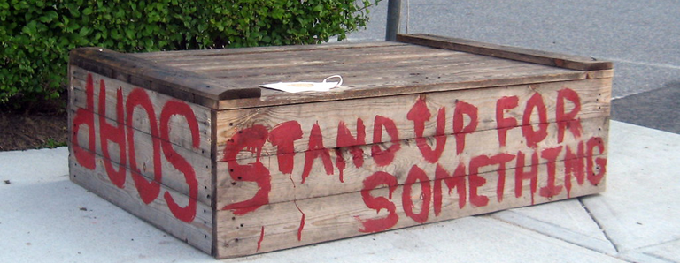 Photo of a large wooden box on the sidewalk, painted on one side with the words "Stand up for something" and 
