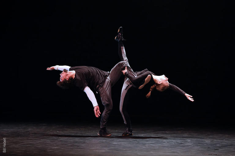 A male and female dancer facing each other both lean back balancing on one leg.
