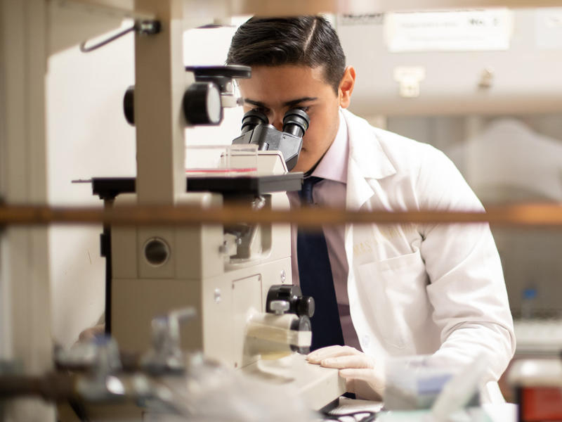 Student in a lab wearing a lab coat and looking into a microscope. 