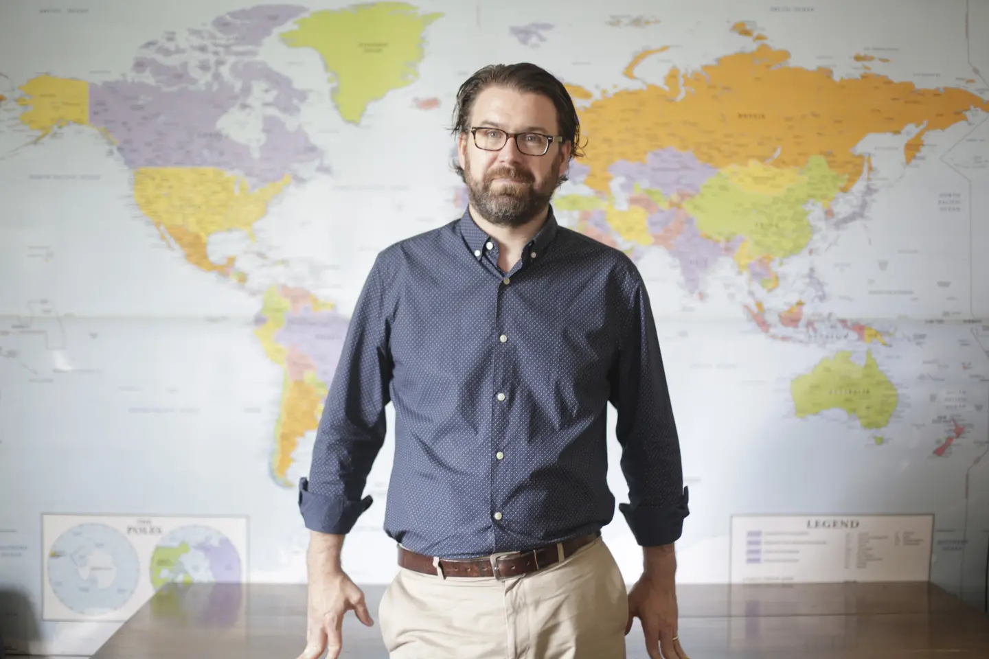 A man leans against a desk with a map of the world behind him