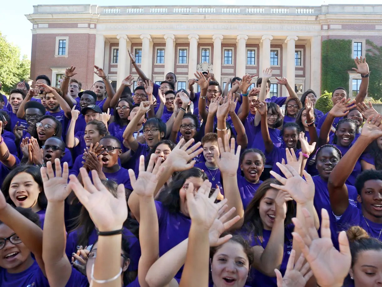 A group of first-generation students at orientation wearing purple shirts and raising their arms.