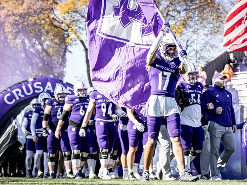 Holy Cross football enters Fitton Field holding a large purple flag with the Holy Cross Athletics shield logo.